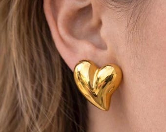 May include: A polished gold-tone heart-shaped stud earring with a smooth, reflective surface. The earring features a puffed, three-dimensional design that sits flush against the earlobe, showcasing a bright, metallic finish and curved edges.