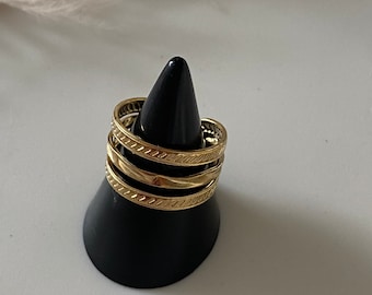 May include: A set of three gold-toned rings stacked on a black conical display stand. The top and bottom rings feature a repeating diagonal etched pattern, while the middle ring has a smooth, polished surface. The rings are metallic and circular.