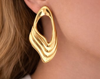May include: A large, gold-toned earring with an irregular, organic shape featuring a smooth, polished surface and a central open cutout. The lower portion of the earring displays a series of three distinct, flowing, wave-like ridges.
