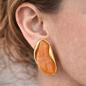 May include: Close-up of a gold-toned earring with an orange, organic-shaped stone. The earring is a clip-on style, with a smooth, polished finish. The design is modern and bold, with a focus on the natural color and shape of the stone.