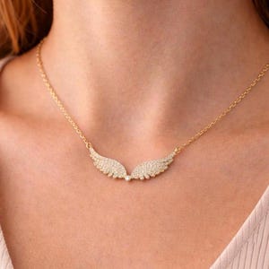 Puede incluir: Un collar dorado con un colgante en forma de dos alas. Las alas est&aacute;n adornadas con peque&ntilde;as piedras brillantes. El collar se lleva alrededor del cuello.