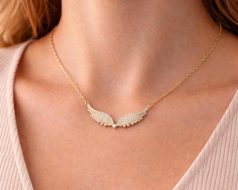 Puede incluir: Un collar dorado con un colgante en forma de dos alas. Las alas est&aacute;n adornadas con peque&ntilde;as piedras brillantes. El collar se lleva alrededor del cuello.