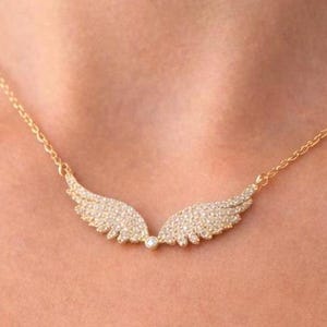 Puede incluir: Un collar dorado con un par de alas de &aacute;ngel, cada una incrustada con peque&ntilde;os cristales brillantes. Las alas est&aacute;n conectadas por una peque&ntilde;a gema redonda. El collar se muestra sobre la piel de una persona.