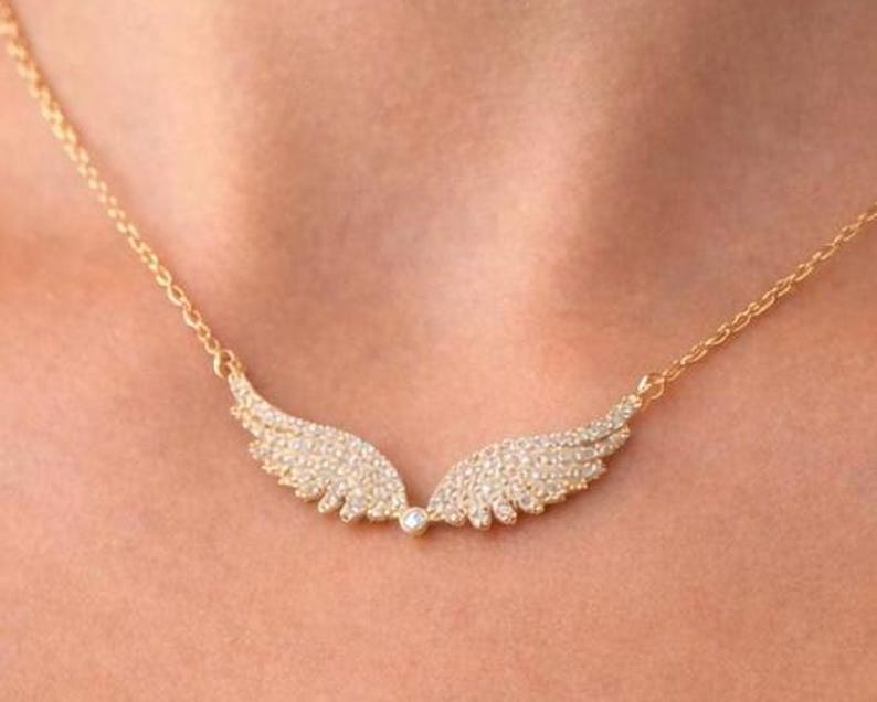 Puede incluir: Un collar dorado con un par de alas de &aacute;ngel, cada una incrustada con peque&ntilde;os cristales brillantes. Las alas est&aacute;n conectadas por una peque&ntilde;a gema redonda. El collar se muestra sobre la piel de una persona.