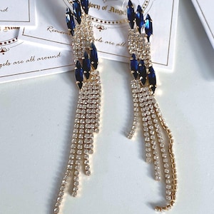 Puede incluir: Un par de aretes largos y colgantes con una cadena dorada y detalles de cristal azul. Los aretes presentan un racimo de cristales azules en la parte superior y una cadena de cristales más pequeños en cascada.
