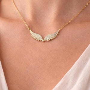 Puede incluir: Un collar dorado con un colgante en forma de alas. Cada ala est&aacute; adornada con peque&ntilde;os cristales brillantes. El collar se lleva alrededor del cuello, descansando sobre el pecho de una persona, contra una parte superior de color claro.