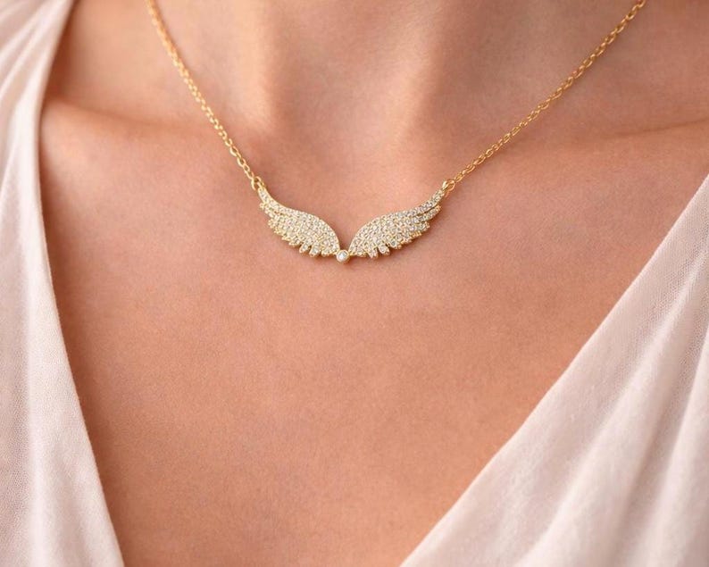 Puede incluir: Un collar dorado con un colgante en forma de alas. Cada ala est&aacute; adornada con peque&ntilde;os cristales brillantes. El collar se lleva alrededor del cuello, descansando sobre el pecho de una persona, contra una parte superior de color claro.