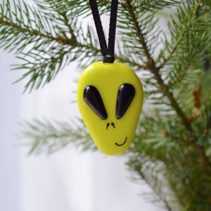 Peut inclure: Ornement en verre jaune en forme de tête d'extraterrestre avec des yeux noirs et un sourire. L'ornement est suspendu à une ficelle noire sur une branche de pin verte.
