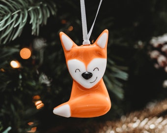 May include: Orange ceramic fox ornament with white accents and a smiling face. The ornament is hanging from a white string on a Christmas tree.