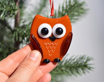 May include: A handcrafted orange glass owl ornament with large white eyes and a black beak. The owl has a red ribbon for hanging and is a festive decoration for a Christmas tree or holiday display.
