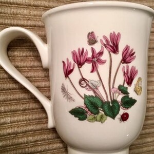 May include: White ceramic mug with a floral design featuring pink cyclamen flowers and green leaves. The mug has a white handle.
