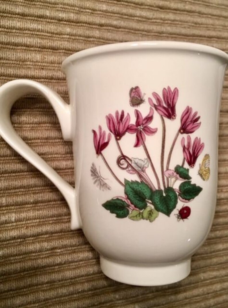 May include: White ceramic mug with a floral design featuring pink cyclamen flowers and green leaves. The mug has a white handle.