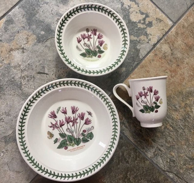 May include: A set of white ceramic dinnerware with a green border and floral pattern. The set includes a large dinner plate, a smaller bowl, and a mug. The floral pattern features pink cyclamen flowers and green leaves.
