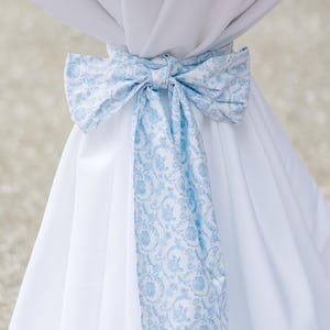 May include: A white fabric bow with a light blue floral pattern, tied around a white draped fabric. The bow is the focal point, with the blue pattern adding a decorative touch to the otherwise plain white fabric.