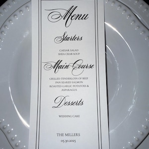May include: A white menu card with black text, featuring elegant script fonts. The menu lists starters, main courses, and desserts, including Caesar salad, grilled tenderloin, and wedding cake. The bottom of the card reads "THE MILLERS 05.30.2025".