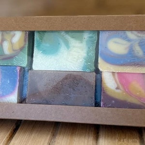 May include: A set of six handmade soap bars in a cardboard box. The soaps are all different colors and have swirls of color throughout. The soaps are arranged in two rows of three.