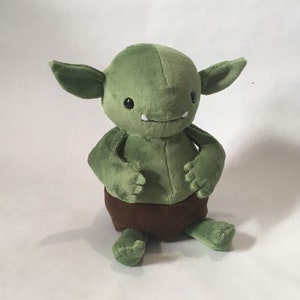 May include: A green plush toy goblin wearing a brown fabric nappy. The goblin has a wide smile and large eyes.