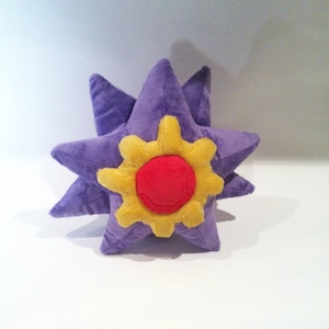 Może przedstawiać: Pluszowa zabawka w kształcie fioletowej gwiazdy z żółto-czerwonym środkiem. Gwiazda ma wiele punktów i miękki, teksturowany wygląd. Zabawka została zaprojektowana tak, aby przypominać postać z popularnej gry wideo.