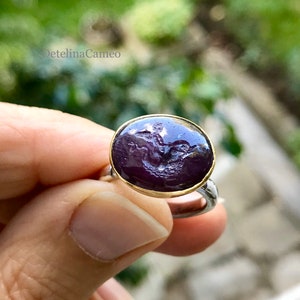 Puede incluir: Un anillo de piedra preciosa púrpura con un engaste de oro. La piedra preciosa está tallada con un diseño detallado de una figura.