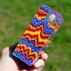 May include: A rectangular block featuring a vibrant, multi-colored pattern of wavy, zigzagging lines in shades of neon orange, yellow, purple, and blue, separated by thin black lines. A small square sticker with a logo and the text stabilizedempire is attached.