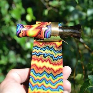 May include: A colorful, patterned smoking pipe with a black mouthpiece and brass accents. The pipe features a vibrant design of swirling rainbow colors and a zig-zag pattern. The background is a blurred view of green foliage.