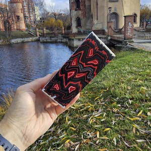 May include: A rectangular decorative item with a striking red, black, and white patterned design. The item is held in front of a scenic background featuring a body of water and a castle-like structure.
