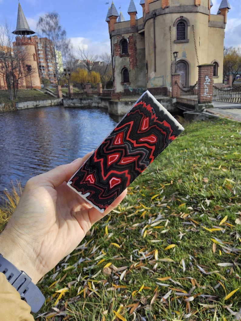May include: A rectangular decorative item with a striking red, black, and white patterned design. The item is held in front of a scenic background featuring a body of water and a castle-like structure.