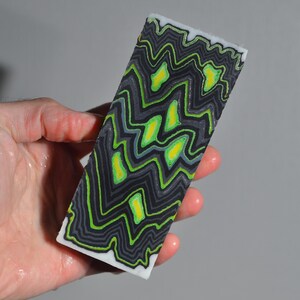 May include: A rectangular block with a black, gray, and neon green zigzag pattern. The design includes yellow accents and a white border. The block is held in a hand.