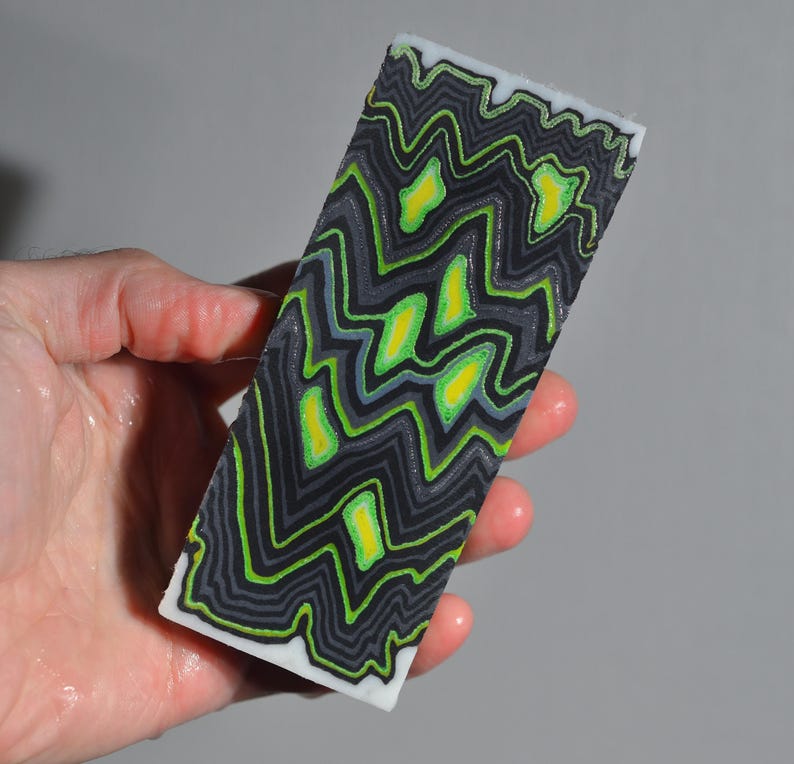 May include: A rectangular block with a black, gray, and neon green zigzag pattern. The design includes yellow accents and a white border. The block is held in a hand.