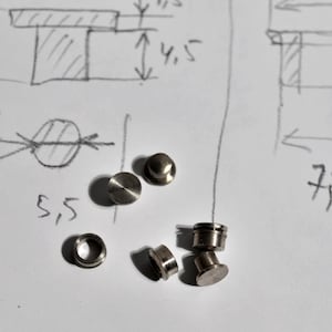 May include: A collection of small, metallic hardware components, including cylindrical and disc-shaped pieces, are scattered on a white surface. Hand-drawn diagrams with measurements in centimeters are visible in the background, suggesting a technical or engineering context.