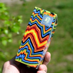 May include: A colorful block of resin with a wavy, abstract pattern in shades of blue, green, yellow, orange, and red. The block has a small, silver sticker with a logo on it.