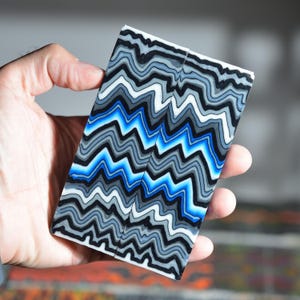 Op de afbeelding: Een rechthoekig blok van composietmateriaal met een herhalend patroon van horizontale, grillige zigzaglijnen in afwisselende tinten zwart, wit, grijs en levendig blauw. Het oppervlak is glad en vlak met een consistent, gelaagd ontwerp.