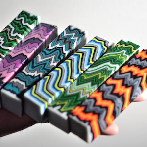 May include: A collection of colorful, rectangular resin blocks with a wavy, zig-zag pattern in various colors. The blocks are held in a hand, showcasing the vibrant designs. Colors include green, purple, teal, blue, yellow, and orange.