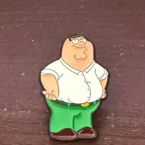 Può includere: Una statuetta di un personaggio dei cartoni animati di un uomo con una camicia bianca, pantaloni verdi e scarpe marroni. Il personaggio ha una corporatura grande, occhiali e un'espressione sorridente. La statuetta è un oggetto da collezione.