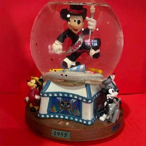 Op de afbeelding: Een Mickey Mouse sneeuwbol met Mickey in een smoking met hoge hoed, die een baton vasthoudt. De basis heeft een circustentontwerp met de jaren 1955 en 1928. De bol heeft confetti binnenin.