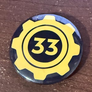 May include: A round button with a yellow cogwheel design around a black circle. Inside the circle is the number 33 in yellow. The button has a dark blue background.