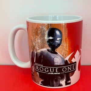Puede incluir: Taza de cerámica blanca con un diseño de la película "Rogue One: A Star Wars Story". La taza muestra un droide gris oscuro sobre un fondo rojo y marrón. Tiene un asa y un interior blancos.