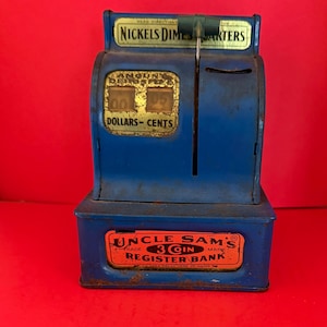 May include: A vintage blue metal Uncle Sam's Register Bank. The bank has a slot for coins and a display showing "00.25". The top reads "Nickels Dimes Quarters". The bottom has the text "Uncle Sam's 3 Coin Register Bank".