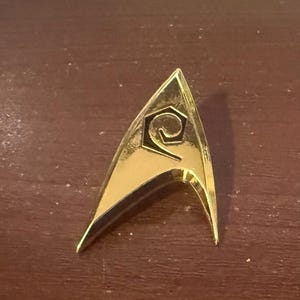 Könnte beinhalten: Ein goldfarbener, dreieckiger Ansteckpin mit einem stilisierten Spiraldesign in der Mitte. Der Pin hat eine polierte, reflektierende Oberfläche und eine klassische, spitze Form. Der Pin ist ein Star Trek-Abzeichen.