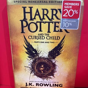 May include: A special rehearsal edition of "Harry Potter and the Cursed Child" book. The cover features a gold and brown color scheme with the title in large, bold letters. A nest with a figure is in the center, with black wings. The book has a red sticker that says "Members Save 20%"