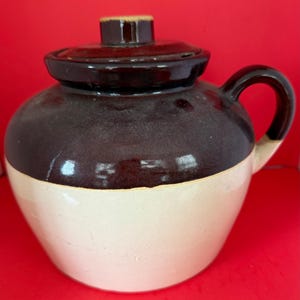 Könnte beinhalten: Ein zweifarbiges Keramikgefäß mit Deckel und Griff. Die obere Hälfte ist dunkelbraun, die untere Hälfte cremefarben. Der Deckel hat einen kleinen, zylindrischen Griff. Das Gefäß hat eine abgerundete Form und einen gebogenen Griff.