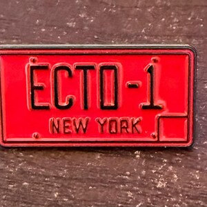 May include: A rectangular red metal pin featuring the text "ECTO-1" and "NEW YORK" in black. The pin resembles a New York license plate. The pin is set against a dark, textured background.