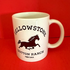 Op de afbeelding: Witte keramische mok met een zwarte paardenafbeelding en de tekst "Yellowstone Dutton Ranch Montana". De mok heeft een gebogen handvat. Op een rode achtergrond.