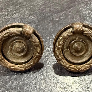 May include: A pair of antique bronze cabinet knobs. Each knob has a circular design with a central recessed area, surrounded by a laurel wreath and a decorative top. The knobs have a weathered, aged appearance.