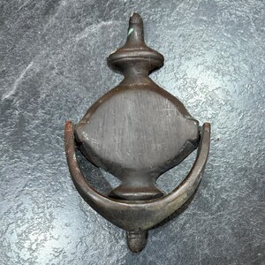 May include: Antique bronze door knocker with a diamond-shaped face and a curved handle. The knocker has a classic design with a decorative top and bottom. The bronze has a weathered appearance, suggesting age and use. A timeless piece for any front door.