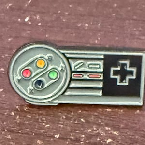 May include: A gray enamel pin shaped like a classic video game controller. The pin features a circular control pad with red, yellow, green, and blue buttons, and a rectangular section with a directional pad.