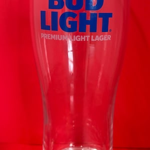 May include: A clear glass beer pint glass with the words "BUD LIGHT" in blue block letters. Below the text is "PREMIUM LIGHT LAGER" in blue. The glass is empty and sits on a red surface.