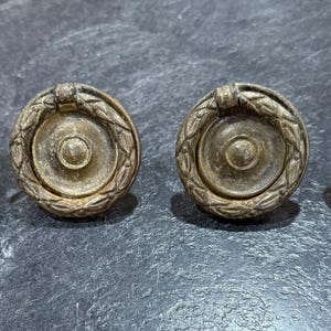 Pode incluir: Quatro puxadores de gaveta antigos em latão com um design circular. Cada puxador apresenta um disco central, uma grinalda circundante e uma pequena pega. Os puxadores estão dispostos em fila sobre uma superfície escura e texturizada.