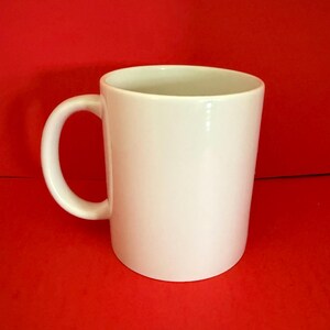 May include: A white ceramic mug with a curved handle. The mug is cylindrical and sits against a solid red background. The mug is empty and appears clean.