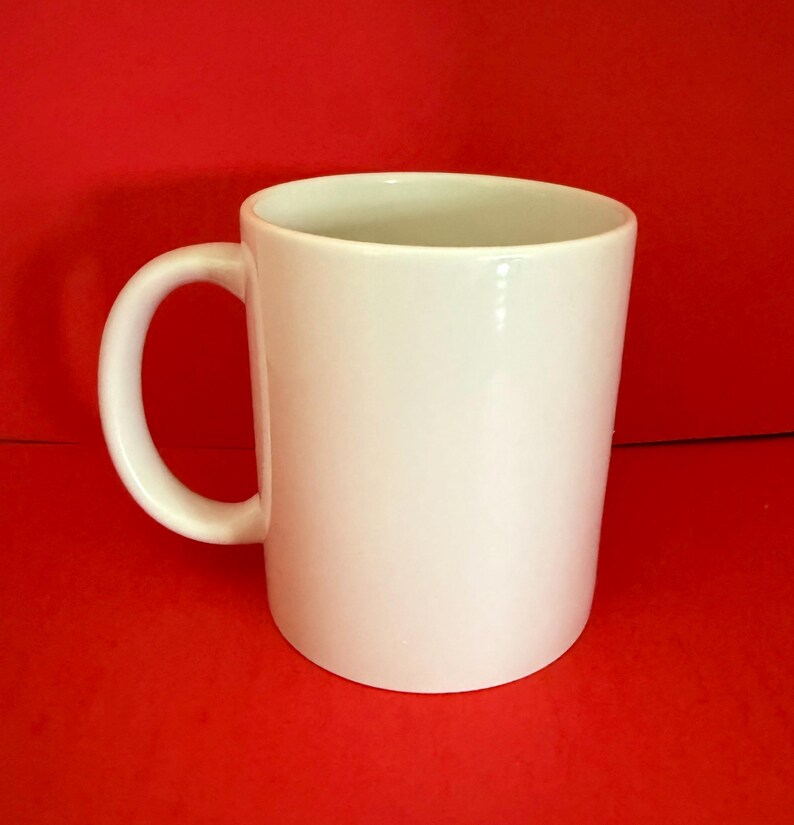 May include: A white ceramic mug with a curved handle. The mug is cylindrical and sits against a solid red background. The mug is empty and appears clean.